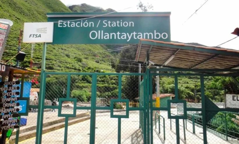 Letrero verde de la Estación Ollantaytambo con rejas de ingreso y montañas verdes al fondo