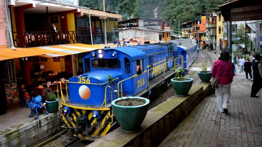 Tren azul de PeruRail transitando por las vías públicas de Machu Picchu Pueblo o Aguas Calientes junto a restaurantes y turistas
